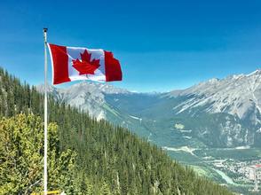 Canadian flag on white pole in foreground with forested hill in background and Rocky Mountains