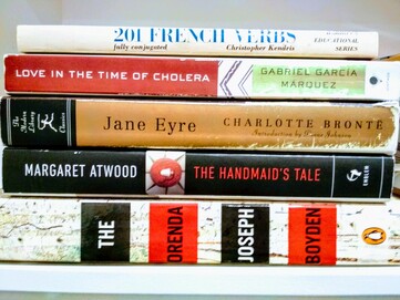 Stack of horizontal books including the Orenda, Handmaid's Tale, Jane Eyre, Love in the Time of Choler, 201 French verbs