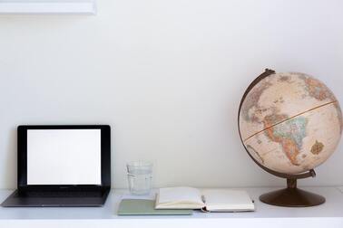 Laptop with blank screen, glass of water, open notebook, and globe on white surface with white background